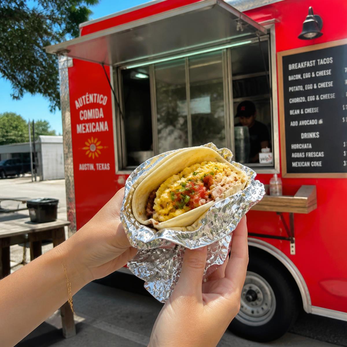 Hands holding a foil-wrapped breakfast taco in front of the red taco truck